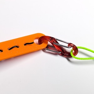 May include: An orange plastic cord organizer with three slots for cords. A red plastic carabiner clip is attached to the organizer and a lime green cord is attached to the carabiner.