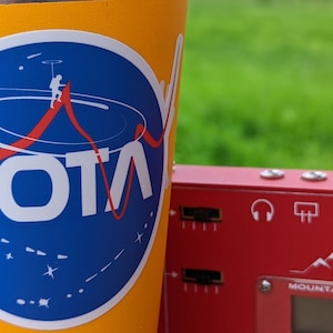 May include: A yellow cup with a blue and white sticker that says "SOTA" and depicts a person climbing a mountain. The cup is next to a red device with the text "Mountain Topper" and a white headphone icon.