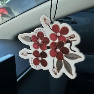 May include: Floral car air freshener featuring red flowers, grey leaves, and white accents. The text "Forbidden Boutique" is printed on the bottom. The air freshener is suspended by a black string.