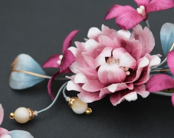 Op de afbeelding: Een close-up van een bloemen haaraccessoire met een grote, meerlagige roze bloem met witte randen, omringd door kleinere magenta bloemen en blauwgroene bladeren. Het ontwerp bevat kleine, bungelende parelachtige kralen.