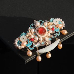 May include: Ornate hair clip with a central red gemstone, surrounded by pearls and smaller red accents. The clip features gold-colored metalwork with turquoise enamel details and dangling peach-colored pearl drops. A white, crescent-shaped element hangs below.