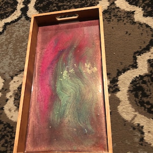 Peut inclure: Un plateau de service en bois avec un dessus en résine transparente. La résine présente un motif tourbillonnant de paillettes roses, vertes et dorées.