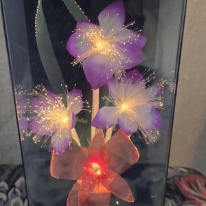 May include: A decorative light fixture with artificial flowers and fibre optic accents. The flowers are purple and orange, with illuminated centres and delicate, glowing fibres. The fixture is encased in a clear, rectangular container.