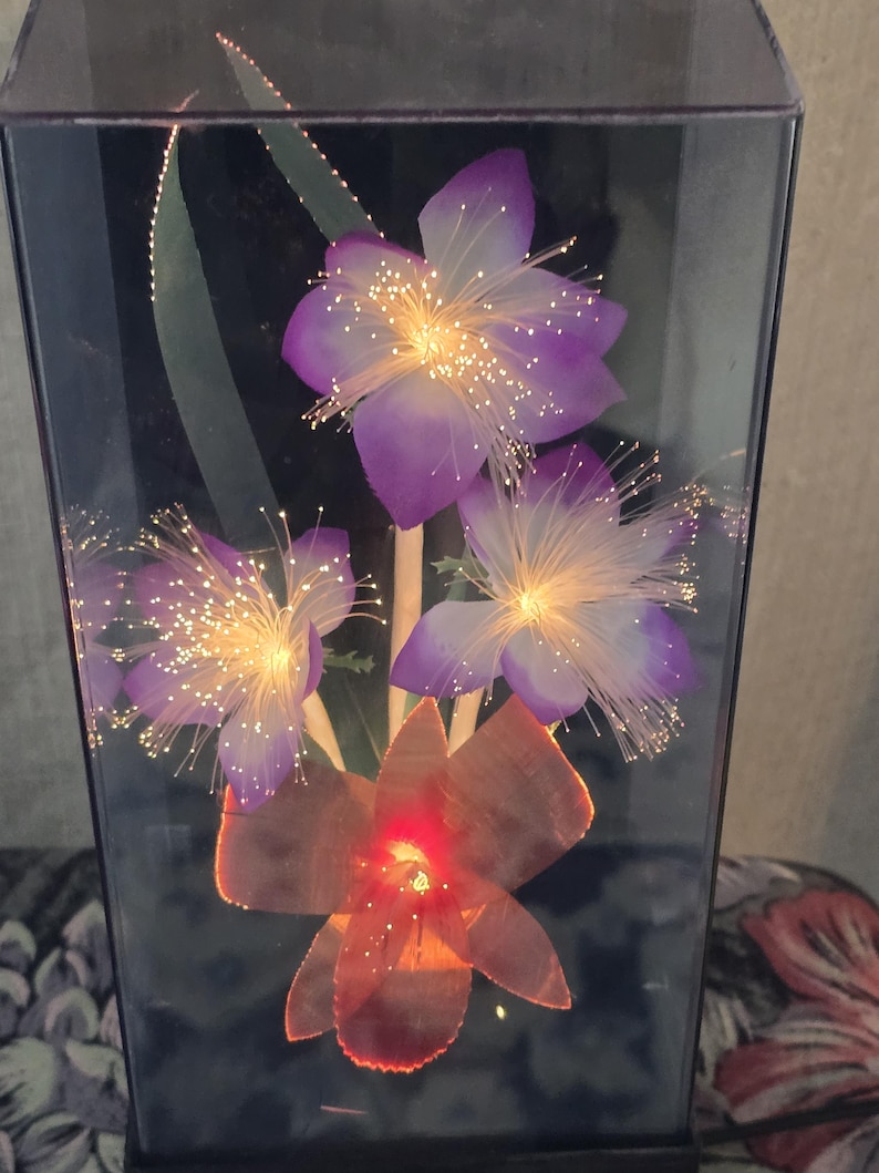 May include: A decorative light fixture with artificial flowers and fibre optic accents. The flowers are purple and orange, with illuminated centres and delicate, glowing fibres. The fixture is encased in a clear, rectangular container.