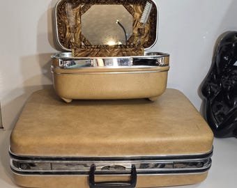 Puede incluir: Maleta de cosméticos vintage de color beige con una maleta más pequeña a juego encima. La maleta superior está abierta, revelando un espejo, un peine y un cepillo. Ambas maletas tienen ribetes plateados y asas negras.