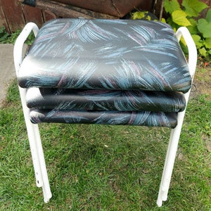 May include: Three folding chairs stacked together, featuring white metal frames. The seats are black, adorned with a pattern of blue, teal, and purple brushstrokes. The chairs are positioned on grass, with a wooden fence and greenery visible.