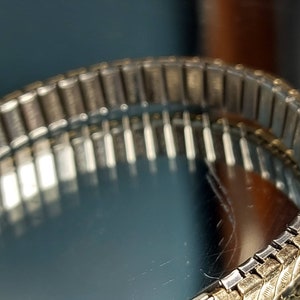 May include: Close-up of a vintage watch band with a silver and gold-toned metal design. The band is flexible and has a repeating pattern of rectangular links. The watch band is on a reflective surface.