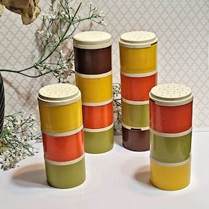 Könnte beinhalten: Vintage Gewürzstreuer-Set in Braun, Gelb, Orange und Grün. Jeder zylindrische Streuer hat einen weißen, perforierten Deckel. Die Streuer sind in Sets von drei oder vier gestapelt und erzeugen einen Retro-Look. Maße in cm.