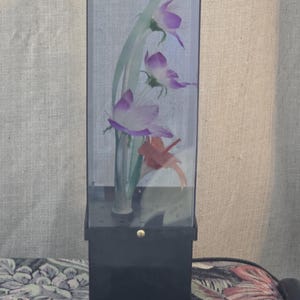 May include: A decorative floral display in a clear rectangular case. The arrangement includes purple and white flowers with green stems and a red accent. The case is set on a black base.