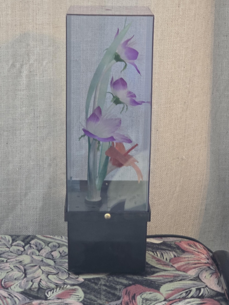 May include: A decorative floral display in a clear rectangular case. The arrangement includes purple and white flowers with green stems and a red accent. The case is set on a black base.