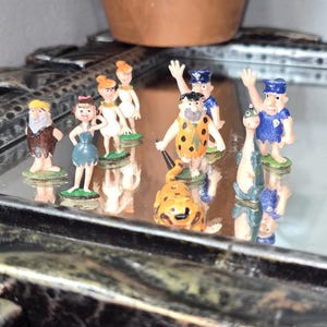 May include: A collection of small, colourful figurines of cartoon characters. The figurines include a man in an orange and black spotted outfit, a woman in a blue dress, and other characters. The figurines are standing on a reflective surface.