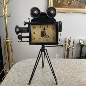 Puede incluir: Un reloj de metal negro y plateado con forma de cámara de cine vintage, montado sobre un trípode. La esfera del reloj tiene un fondo color crema con la palabra "photography" y una imagen de cámara vintage. El reloj está sobre un fondo gris claro.