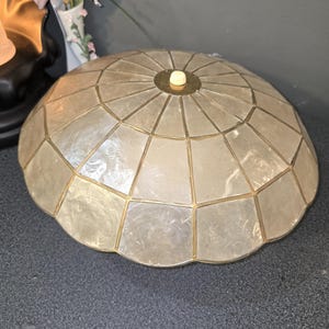Puede incluir: Una pantalla de lámpara en forma de cúpula construida con numerosos paneles de concha translúcidos e iridiscentes dispuestos en un patrón radial. Los paneles están unidos por finas tiras de metal dorado y un pequeño remate circular blanco.