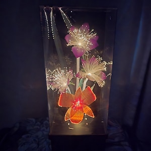May include: A decorative light fixture with illuminated artificial flowers in a clear rectangular container. The flowers are purple and orange with fiber optic strands that emit a soft glow. The base is black.