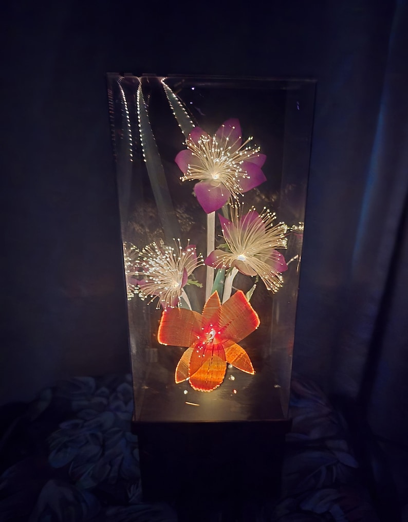 May include: A decorative light fixture with illuminated artificial flowers in a clear rectangular container. The flowers are purple and orange with fiber optic strands that emit a soft glow. The base is black.