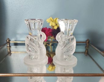 May include: Two clear glass candle holders with swan designs. The candle holders have a frosted base and a clear, faceted top. The swans have detailed wing and neck features. The candle holders are displayed on a mirrored surface.