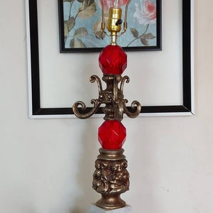 Peut inclure: Une lampe de table vintage avec une base et des accents dorés, avec deux sphères rouges en forme de cristal. La lampe a un design décoratif et une ampoule transparente. Deux petites plantes succulentes en pot se trouvent à gauche de la lampe.