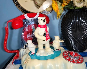 May include: A vintage Coca-Cola telephone featuring a red handset and a base with polar bear figurines. The base has a dial pad and a small ice floe with three polar bears. The bears are holding Coca-Cola bottles and wearing red scarves.