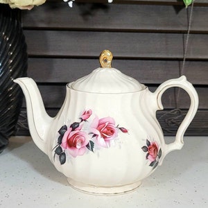 May include: Cream-colored porcelain teapot with a lid, featuring pink rose floral designs and gold trim. The teapot has a curved spout and handle, and a gold-colored knob on the lid. The teapot is sitting on a white surface.