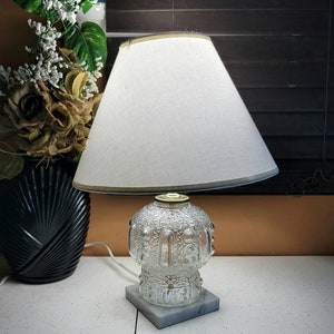 Op de afbeelding: Een heldere glazen tafellamp met een witte lampenkap en een grijze marmeren voet. De lamp heeft een decoratieve, patroon glazen behuizing en een goudkleurige metalen accent aan de bovenkant. De lampenkap is afgewerkt met een gouden rand.