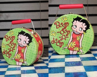 May include: A round, green metal tin with a red handle and a cartoon image of Betty Boop. The tin features the phrase "Boop-Oop-A-Doop" in a whimsical font, along with floral accents. The sides are red with floral patterns.