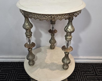 May include: A vintage two-tiered side table with a cream-colored marble top and a lower shelf. The table features ornate, gold-toned metal legs with clear crystal accents. The top has a decorative, filigree edge.