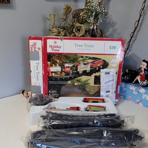 May include: A boxed Holiday Time Tree Train set. The box is red and white with the words "Holiday Time" and "Tree Train" printed on it. The set includes a train, tracks, and other accessories. The train is red, green, and silver.