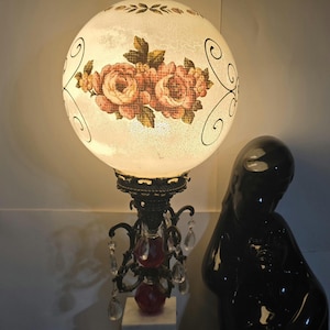 May include: A vintage table lamp with a round, frosted glass shade decorated with pink and peach floral patterns. The lamp has a dark metal base with crystal droplets and a decorative finial. A black statue is in the background.