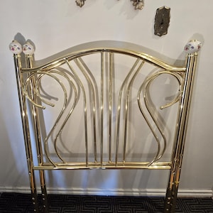 May include: A gold-colored metal headboard with a curved design. The headboard has vertical bars and decorative swirls. The top of the headboard has two decorative ceramic finials with floral patterns.