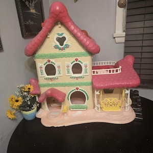 Può includere: Una casa delle bambole vintage in plastica dai colori pastello. Il tetto è rosa e le pareti sono beige con accenti verdi e rossi. Include finestre a forma di cuore, un'altalena e una piccola campana. Un vaso di fiori gialli è nelle vicinanze.