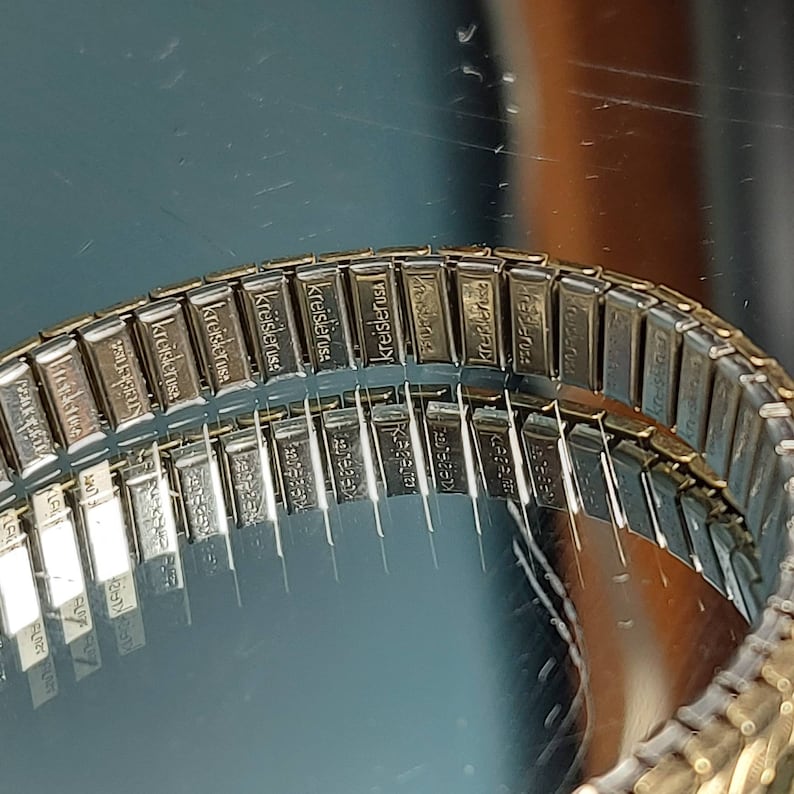 May include: Close-up of a vintage Kreisler USA watch band. The band is made of small, rectangular metal links, each stamped with the brand name. The band is gold-toned and reflects the surrounding light, creating a shiny appearance.