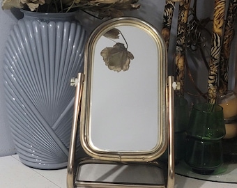 May include: A tabletop vanity mirror with a polished gold-tone metal frame and an arched top. The mirror is mounted on a swivelling stand with two side knobs that allow for tilting the glass. The base consists of a simple metal bar structure.