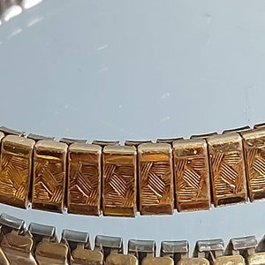 May include: Close-up of two vintage watch bands. The top band is gold-toned with a repeating rectangular pattern. The bottom band is silver-toned. Both bands are flexible and designed to fit a wrist.