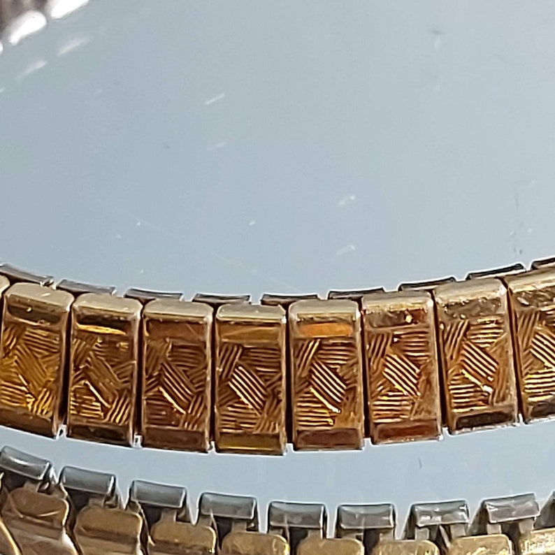 May include: Close-up of two vintage watch bands. The top band is gold-toned with a repeating rectangular pattern. The bottom band is silver-toned. Both bands are flexible and designed to fit a wrist.