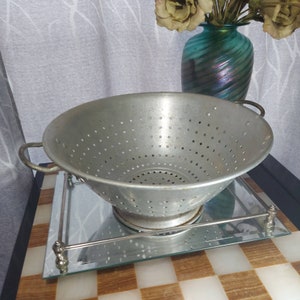 May include: A vintage, aluminum colander with numerous small holes, featuring two side handles. The colander sits on a mirrored tray with a decorative metal frame. A turquoise vase with flowers is in the background. The colander is a kitchen utensil.