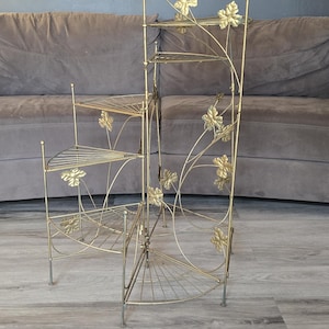 May include: A gold-toned metal plant stand featuring a multi-tiered, corner-fitting design with four semi-circular shelves. The frame is decorated with thin metal vines and leaf-shaped accents, supported by slender legs with a wire-grid shelf structure.
