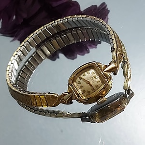 May include: Vintage Gruen Veri-Thin watch with a gold-tone case and a stretch band. The watch face is cream-colored with gold numerals and hands. The band is a mix of gold and silver-tone metal.