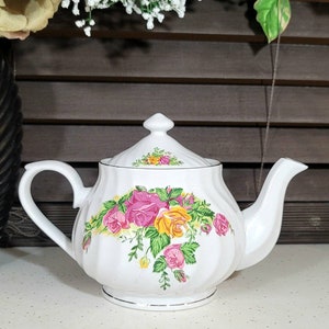 May include: White ceramic teapot with a floral design of pink, yellow, and green roses. The teapot has a curved handle, a spout, and a lid with a small knob. Silver trim accents the base and lid. A classic design for tea.