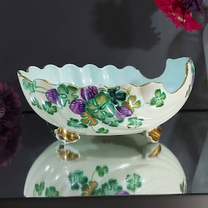 May include: Cream-colored, oval-shaped ceramic bowl with a scalloped rim and three gold-colored feet. The bowl features a light blue interior and is decorated with green, purple, and gold floral and clover designs.
