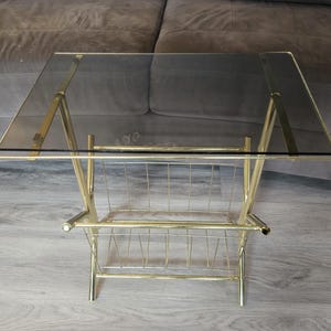 May include: A rectangular side table featuring a polished gold-tone metal frame and a dark, semi-transparent smoked glass top. The base includes an integrated wire rack for storage, constructed with thin vertical metal rods between the legs.