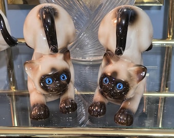 May include: Two ceramic Siamese cat figurines. The cats have distinctive blue eyes and brown markings. They are posed with their tails raised, and their bodies arched. The figurines are displayed on a glass shelf. The finish is glossy.