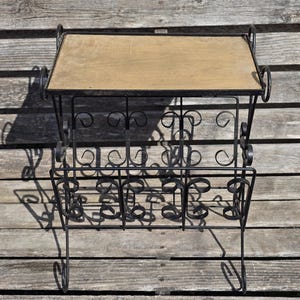 Peut inclure: Un porte-revues en métal noir avec un dessus en bois. Le porte-revues présente un design décoratif en volutes et une section inférieure pour ranger les magazines. Le porte-revues est sur une surface en bois.