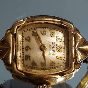 May include: Close-up of a vintage gold-toned Gruen Veri-Thin wristwatch. The watch face is rectangular with gold numerals and hands. The words "SWITZERLAND" and "GRUEN Veri-Thin" are visible. The watch has a gold band.