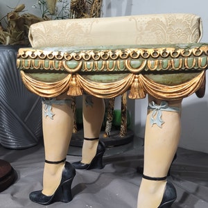Op de afbeelding: Een decoratieve kruk met poten in de vorm van vrouwenbenen met zwarte sleehakken. De kruk heeft een lichtgroene en gouden afwerking, een patroon kussen en lichtblauwe strikken op de benen. Een uniek meubelstuk.