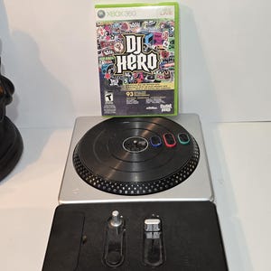 Peut inclure: Un boîtier de jeu vidéo DJ Hero pour Xbox 360 avec une couverture au style graffiti coloré, placé derrière une platine contrôleur noire et argentée. Le contrôleur comprend un plateau circulaire avec trois boutons colorés et une base noire avec deux curseurs.