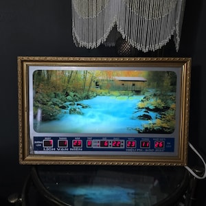 Op de afbeelding: Een goudomrand digitale klok met een rivierlandschap met een overdekte brug. De klok toont de datum, tijd en temperatuur in rode digitale cijfers. De tekst "LỊCH VẠN NIÊN" is zichtbaar.