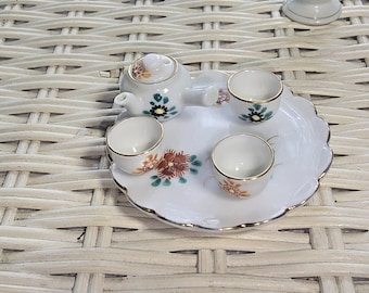 May include: A miniature white porcelain tea set with a teapot, three teacups, and a serving tray. The set is decorated with floral patterns in shades of green, brown, and orange, and has gold trim. The tray has a scalloped edge.