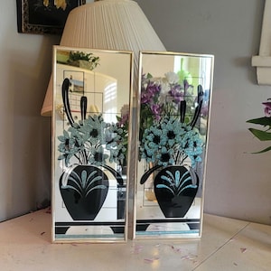 May include: A pair of rectangular wall mirrors with thin gold-tone metal frames. Each mirror features a screen-printed graphic of a black vase containing a bouquet of teal flowers, accented by black geometric lines and a black base design.