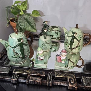 Puede incluir: Un juego de té vintage con un diseño verde claro de vetas de madera. El juego incluye una tetera, azucarero, lechera y dos figuras. Cada pieza presenta una figura verde oscuro y detalles marrones. El juego se exhibe en una bandeja con espejo.