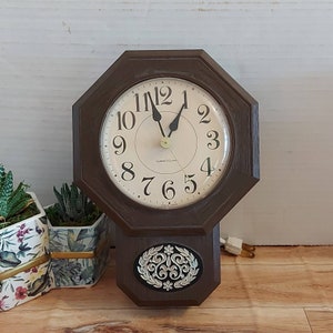Puede incluir: Un reloj de pared octogonal marrón con una esfera crema y números negros. El reloj tiene una sección inferior decorativa con un diseño floral plateado y negro. La esfera del reloj dice "Westclox".